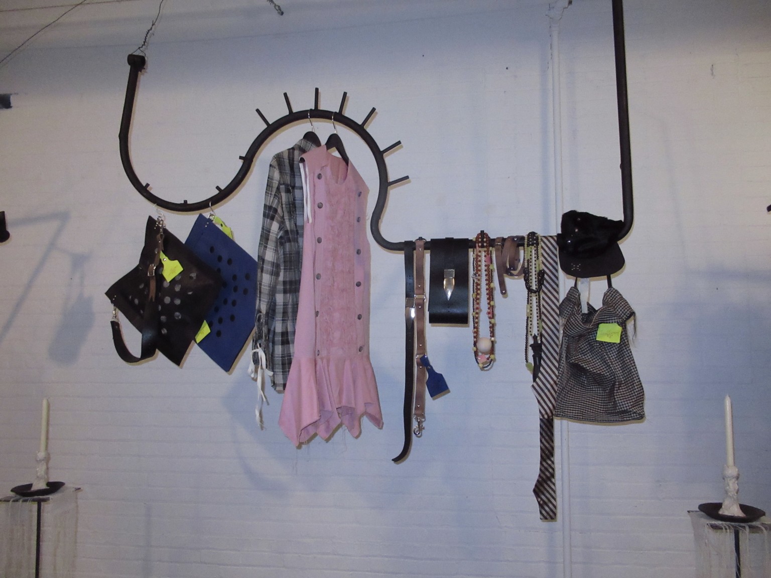 Clothes, bags, belts and necklaces hang from a curved black display rail against a white brick wall, with candles below.