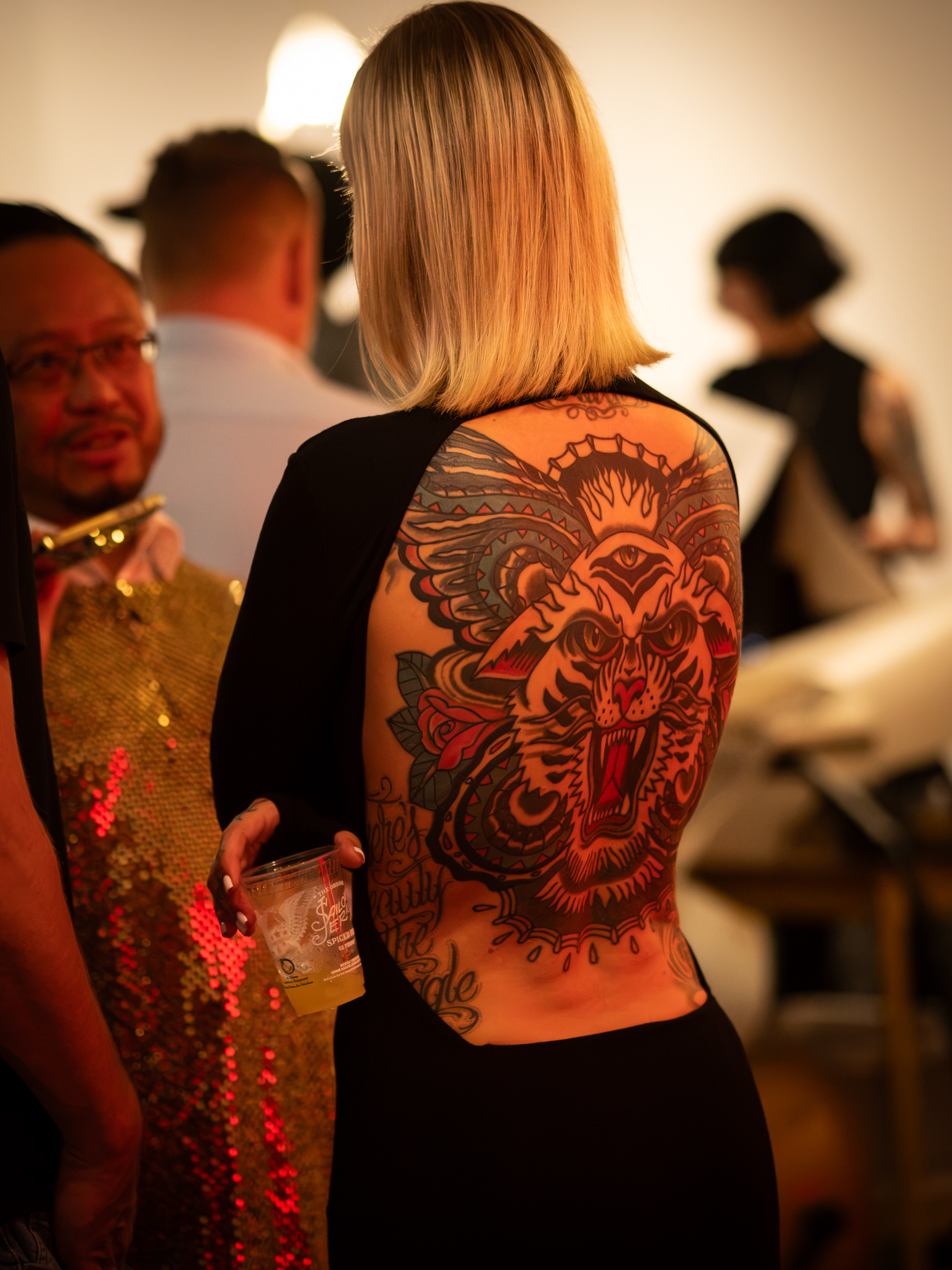 A person holding a drink wears a black outfit with an open back revealing a large tiger tattoo, while talking with another guest.