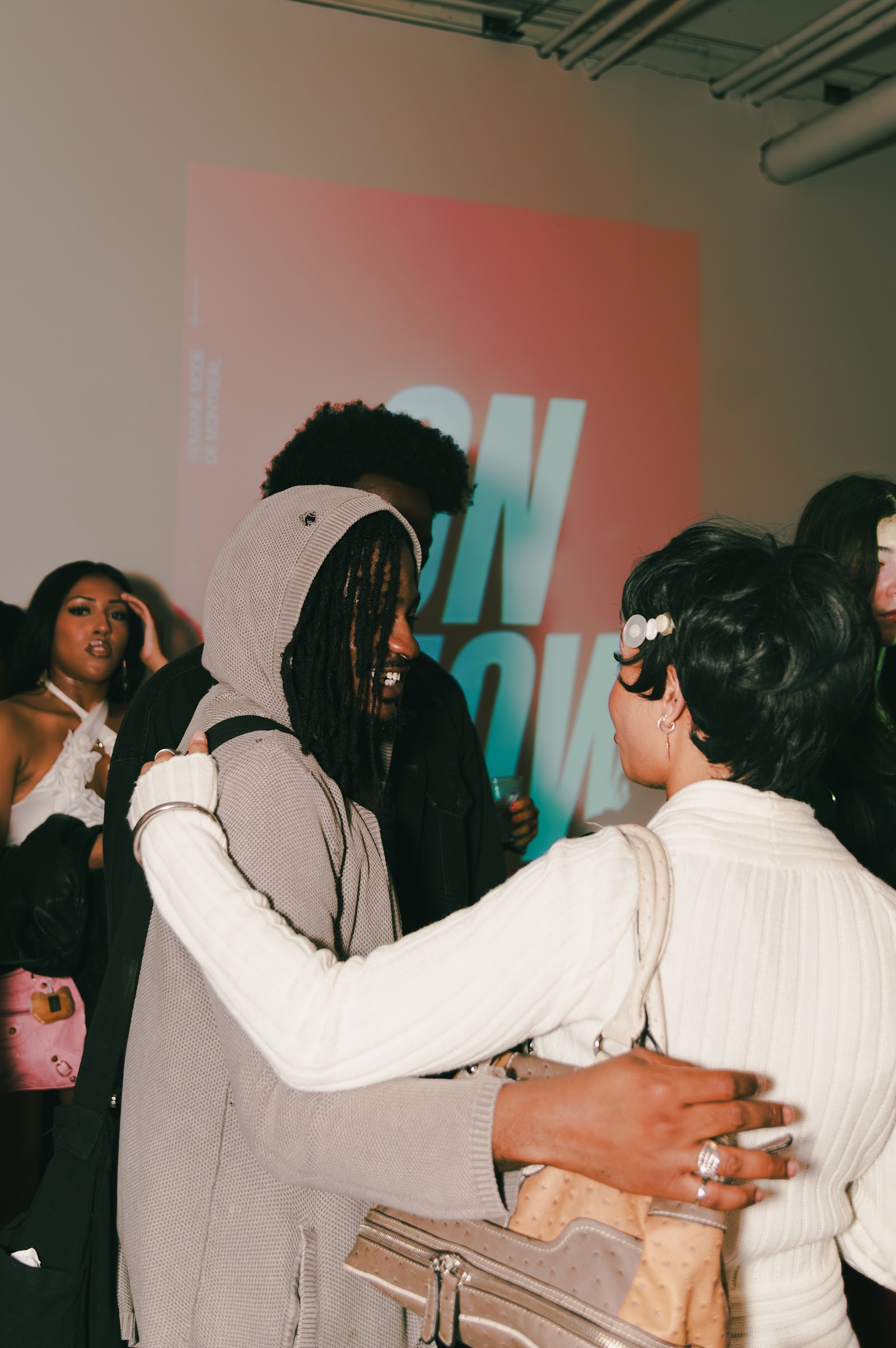 People gather in an embrace in front of the projected ON SHOW title at Atelier Textile.