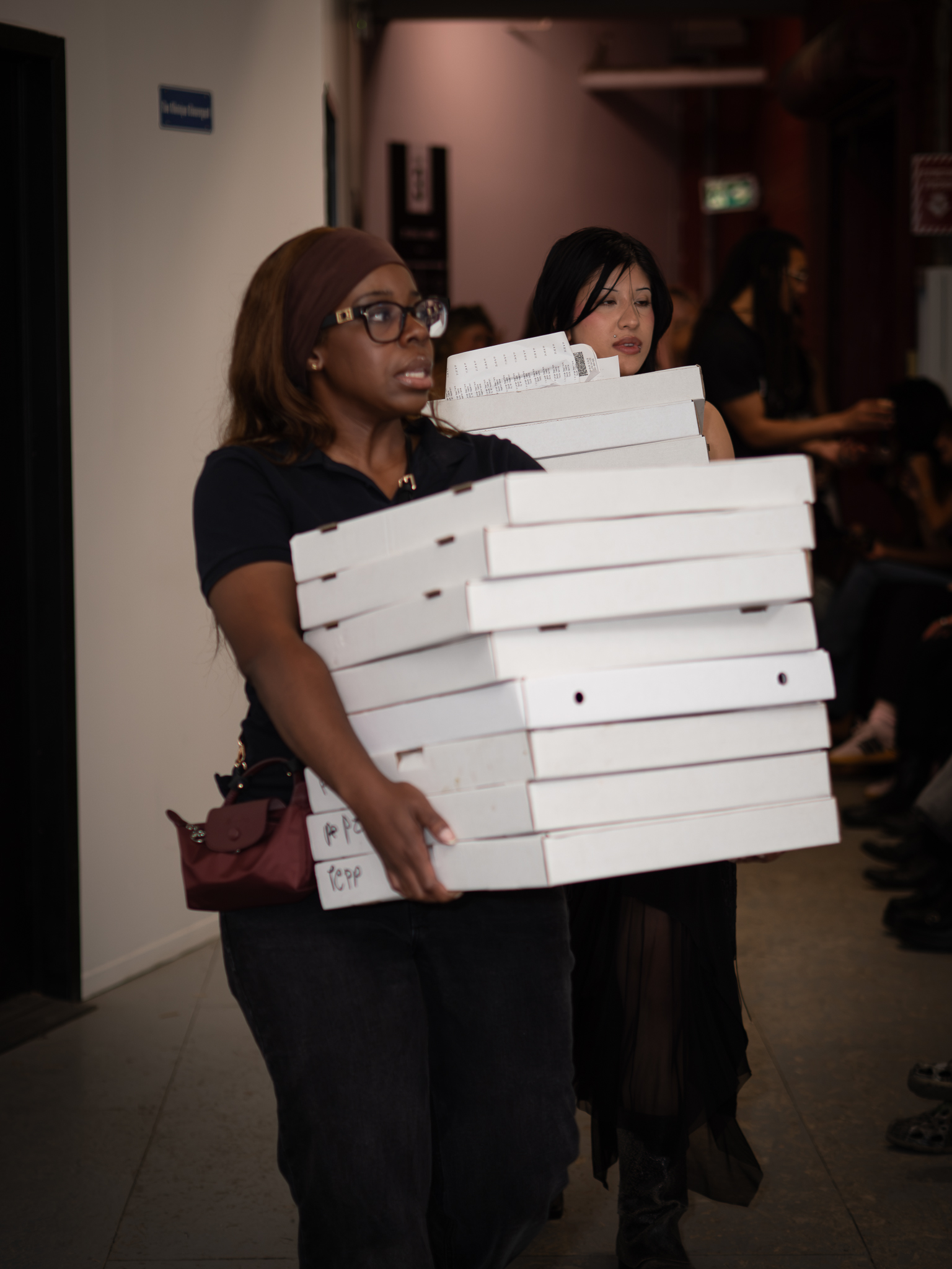 A person carries a stack of white pizza boxes through the doorway, with another person just behind.