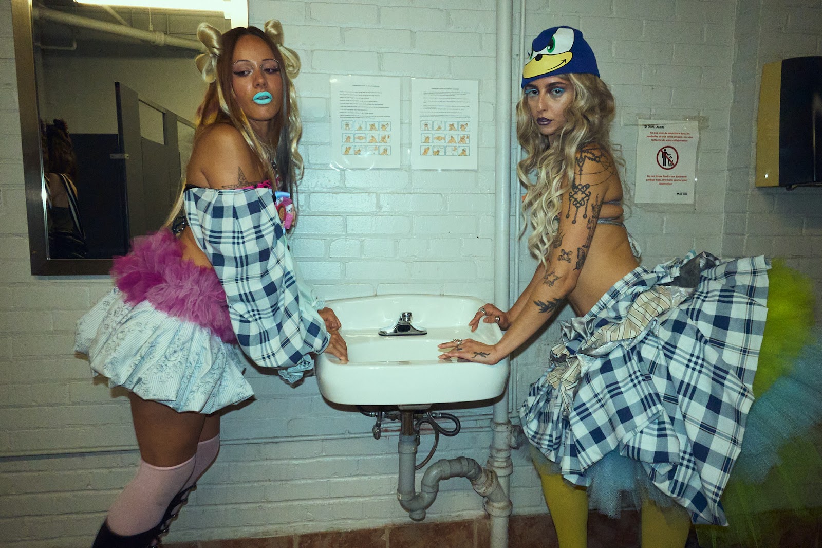 Two models in colourful checked and layered outfits pose on either side of a white bathroom sink against white brick walls.