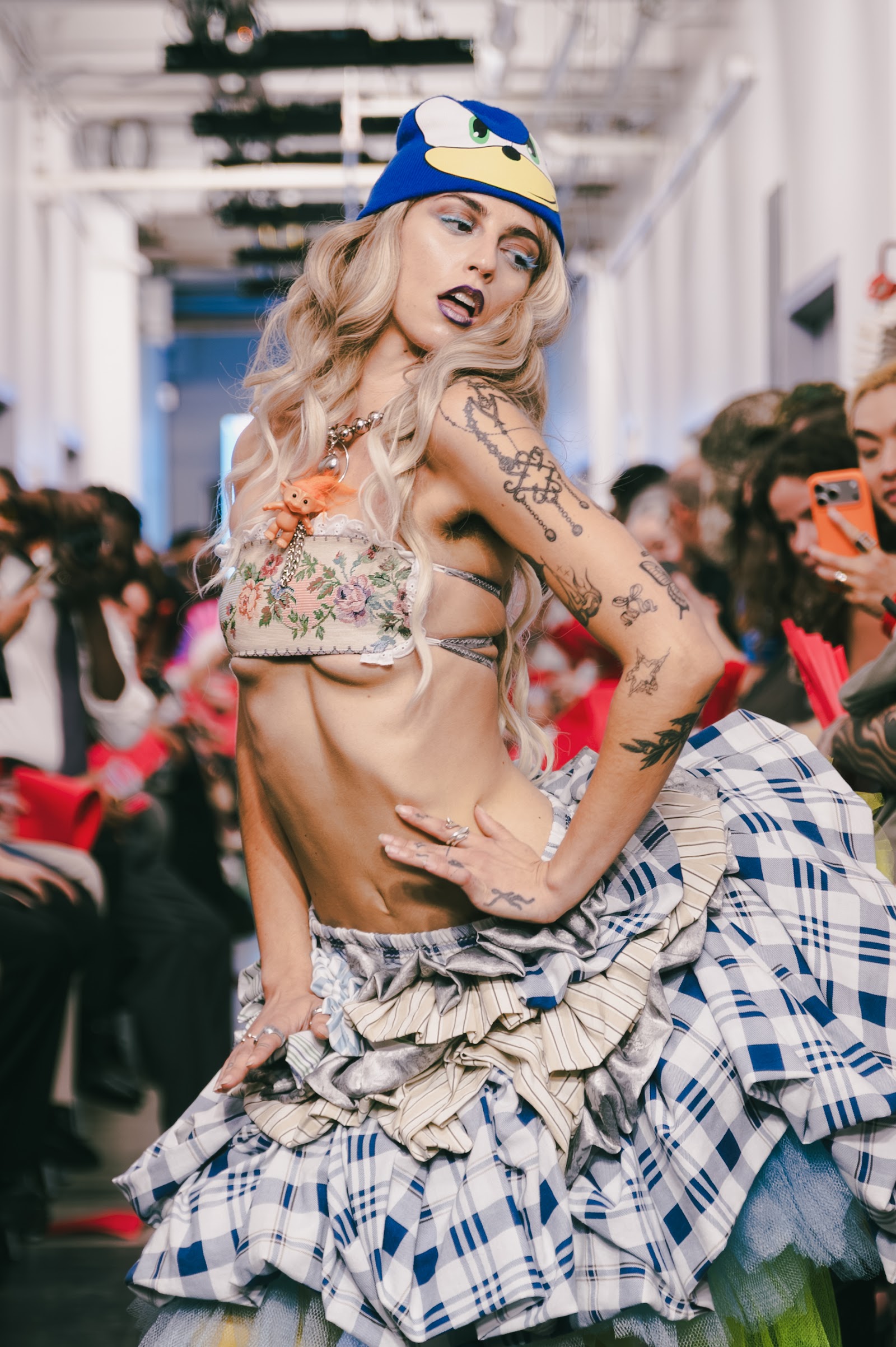 A tattooed model in a blue cartoon hat, floral cropped top and layered checked skirt tilts their head towards the camera on the runway.