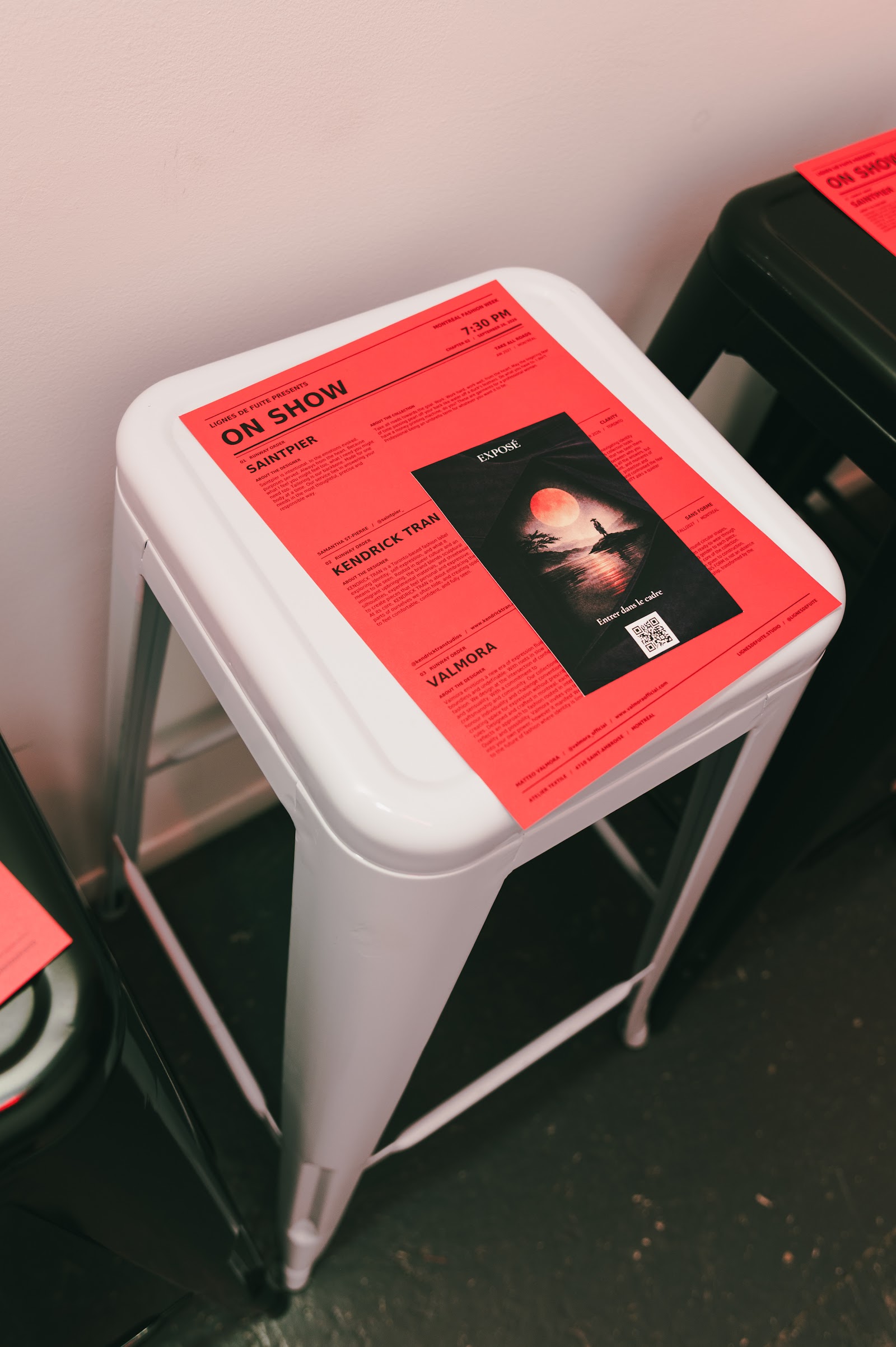 A red ON SHOW programme rests on a white stool beside a black stool in the corridor at Atelier Textile.