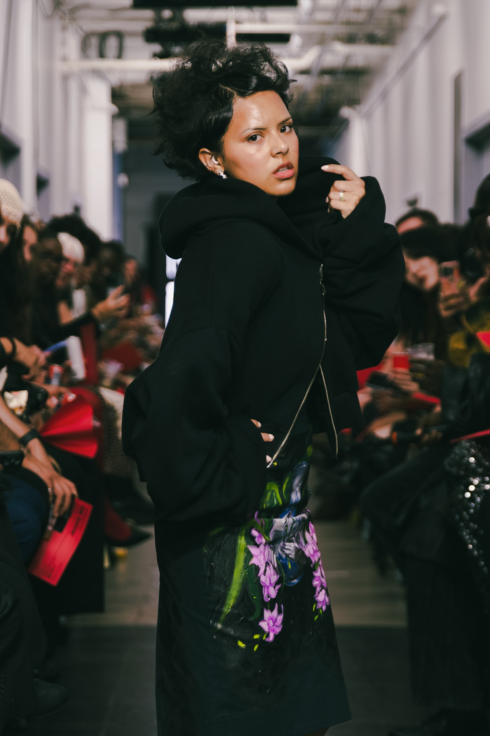 A model turns towards the camera, lifting the collar of a voluminous black jacket above a dark garment with purple floral details.