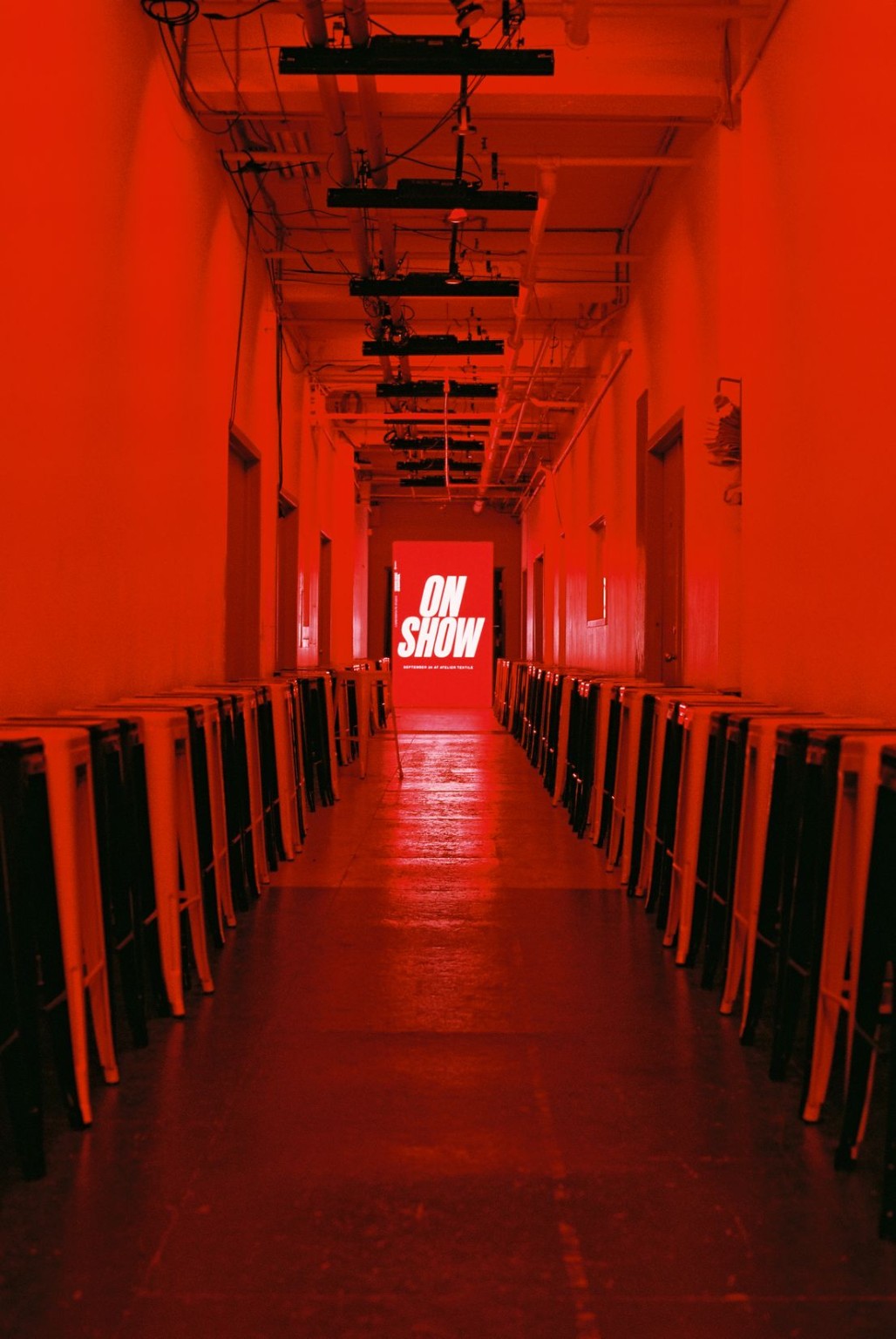 An empty corridor glows red, with two rows of stools facing a screen displaying ON SHOW and overhead lighting rigs.