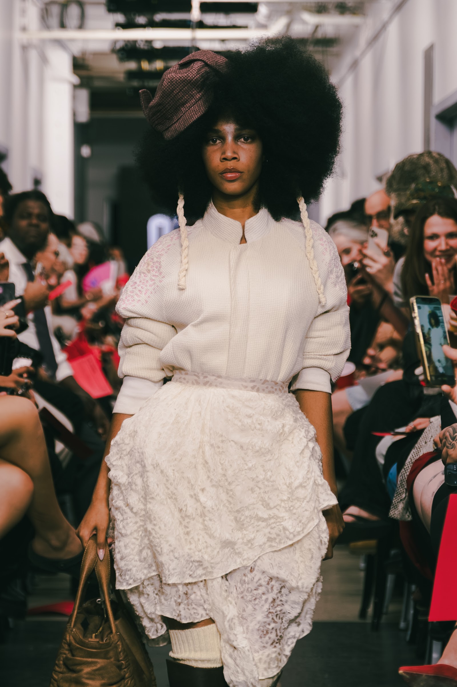 A model wears a white ribbed top, layered white lace skirt, large textured hairstyle and small brown hat on the runway.