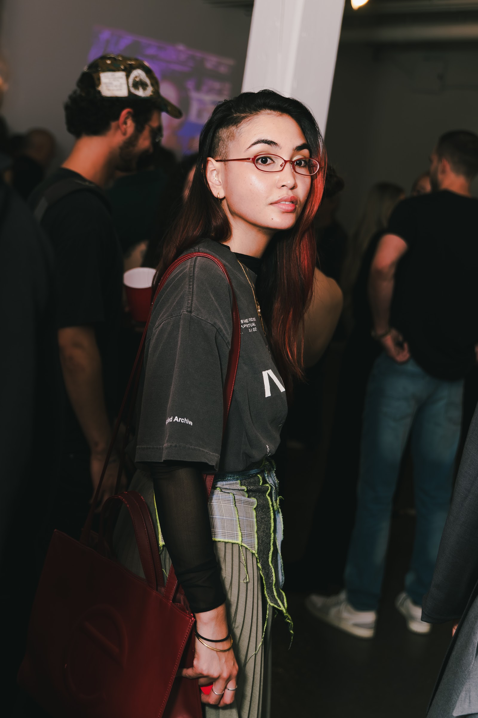 A guest wearing red glasses and a dark T-shirt turns towards the camera, holding a brown bag among people in the room.
