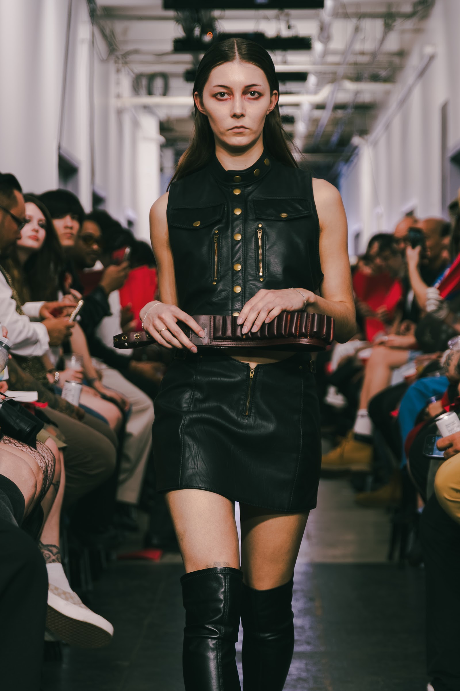 A model wears a black leather vest with brass fastenings, a short black skirt and tall boots, carrying a structured brown bag.