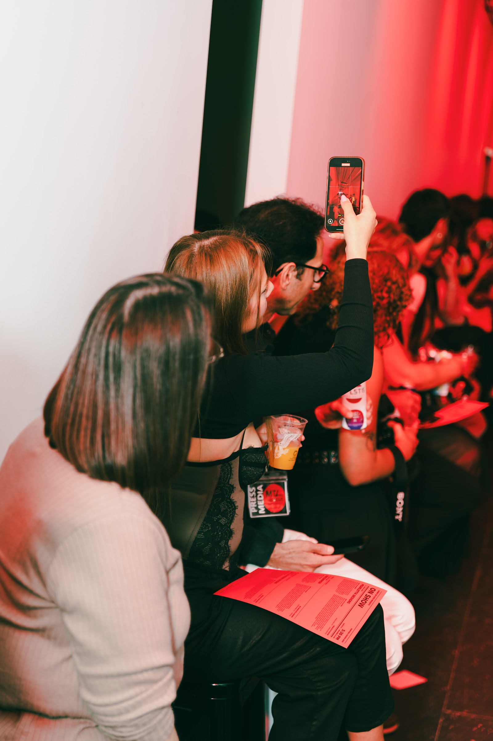 Guests sit closely together under red light, holding drinks and red programmes; one raises a phone to photograph the runway.