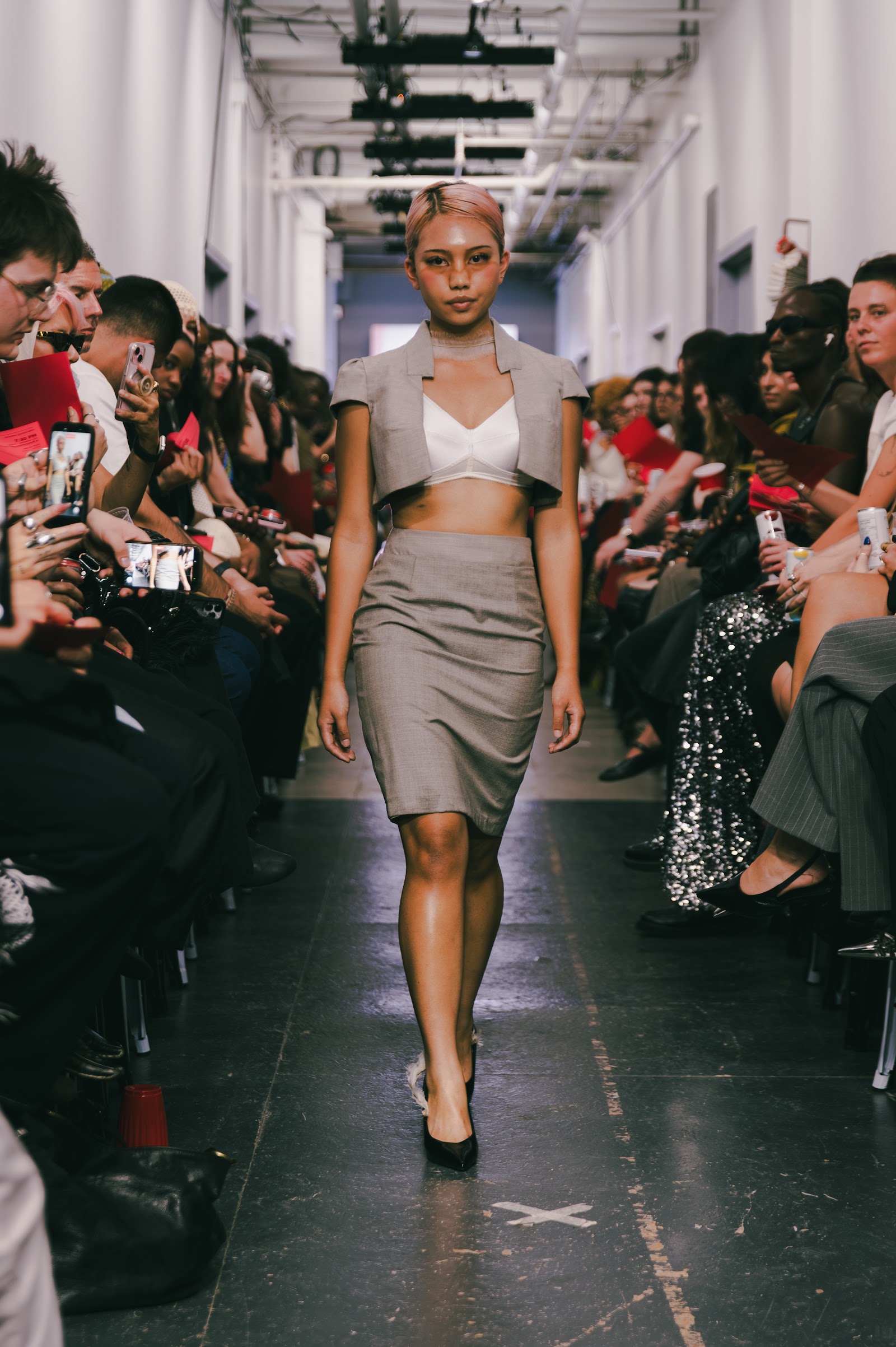A model walks in a grey cropped tailored vest, white top and grey knee-length skirt, with spectators seated along both walls.