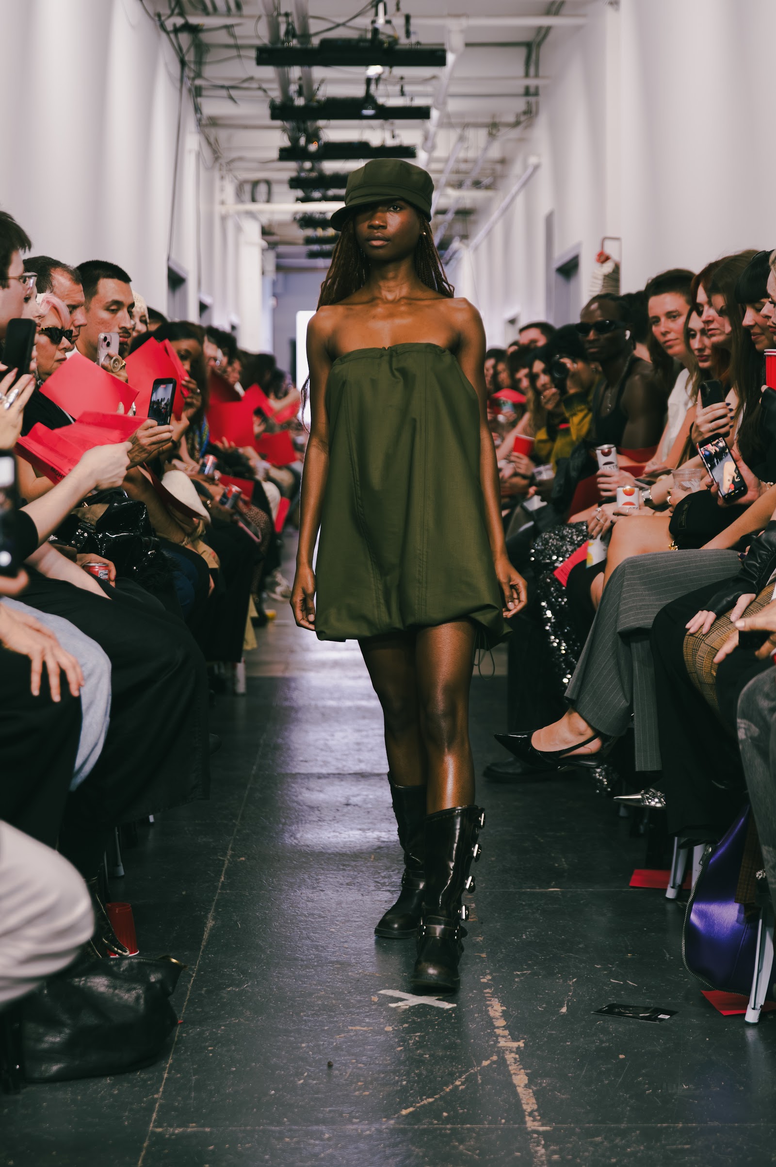 A model in an olive strapless rounded mini dress, matching hat and black boots walks between closely seated guests.