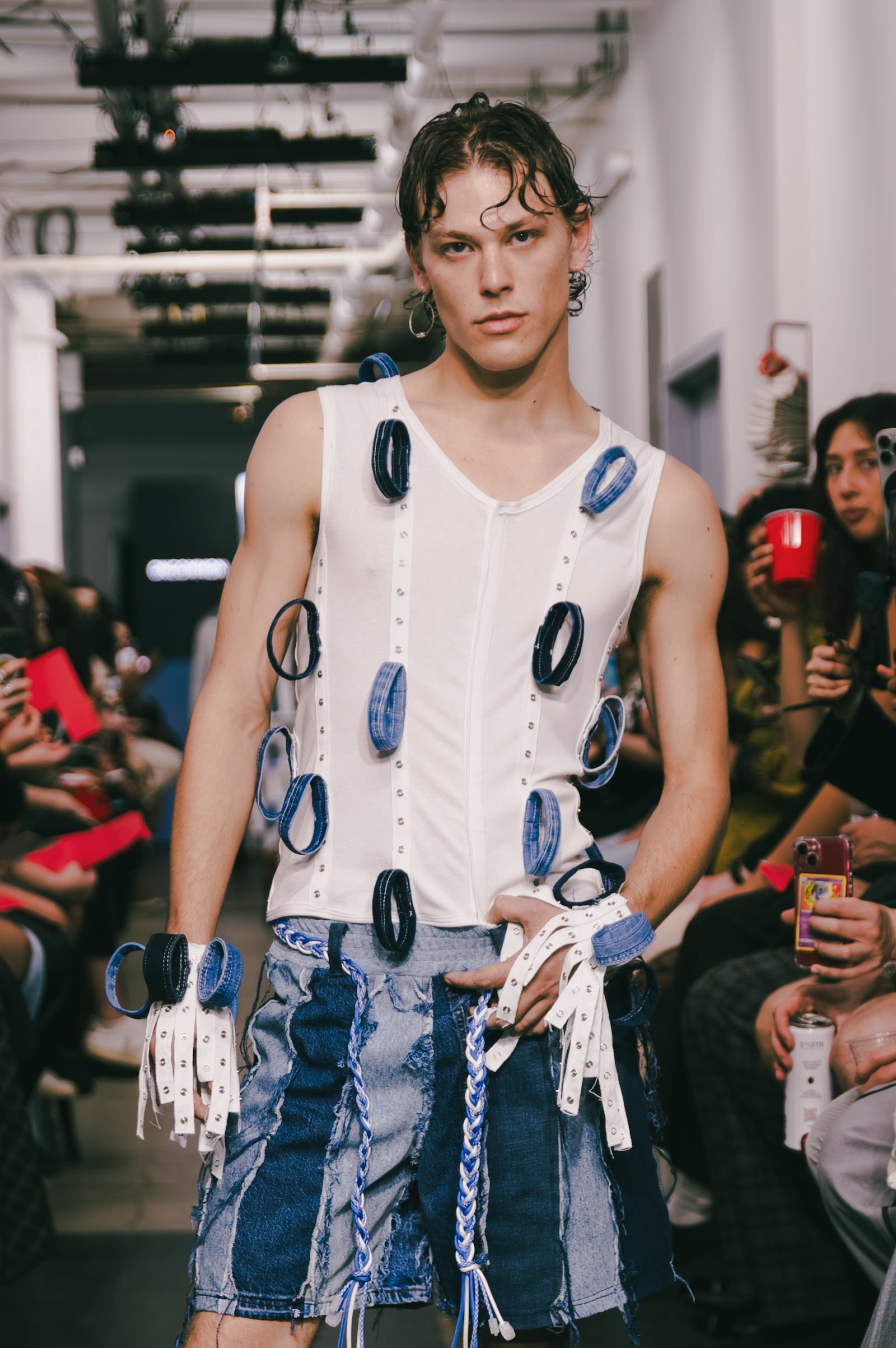 A model wears a white sleeveless top with blue loops and hanging fabric strips above a pieced denim skirt on the runway.
