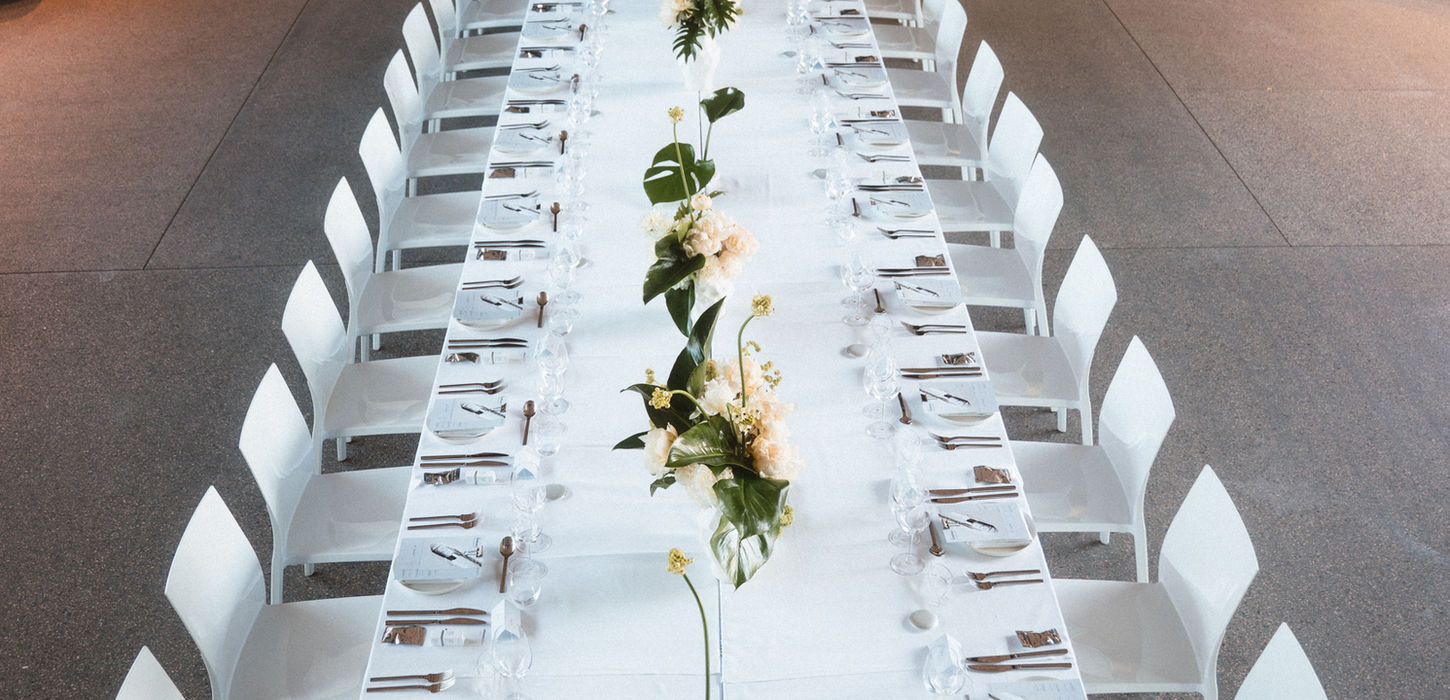 Long dining table with white chairs, elegantly set with white linens, glassware, and floral centerpieces. Wall reads “The Ordinary” in minimalist decor.