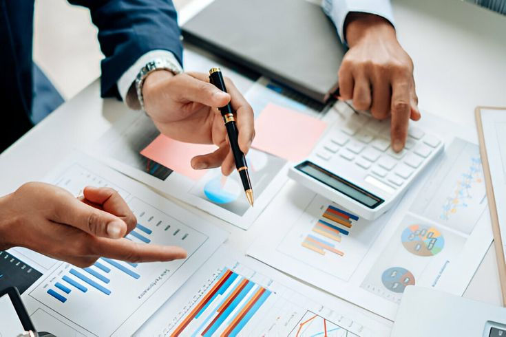 Hands in business suits point at printed charts and a calculator on a desk, analyzing colorful graphs in a meeting.