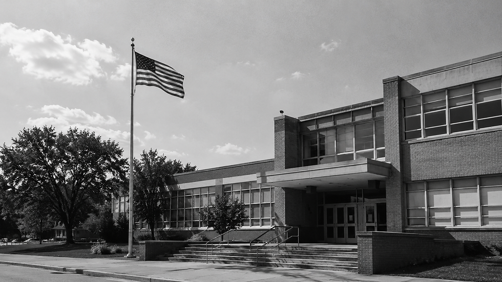 Black-and-white photo of a school building with an American flag waving outside under a cloudy sky.