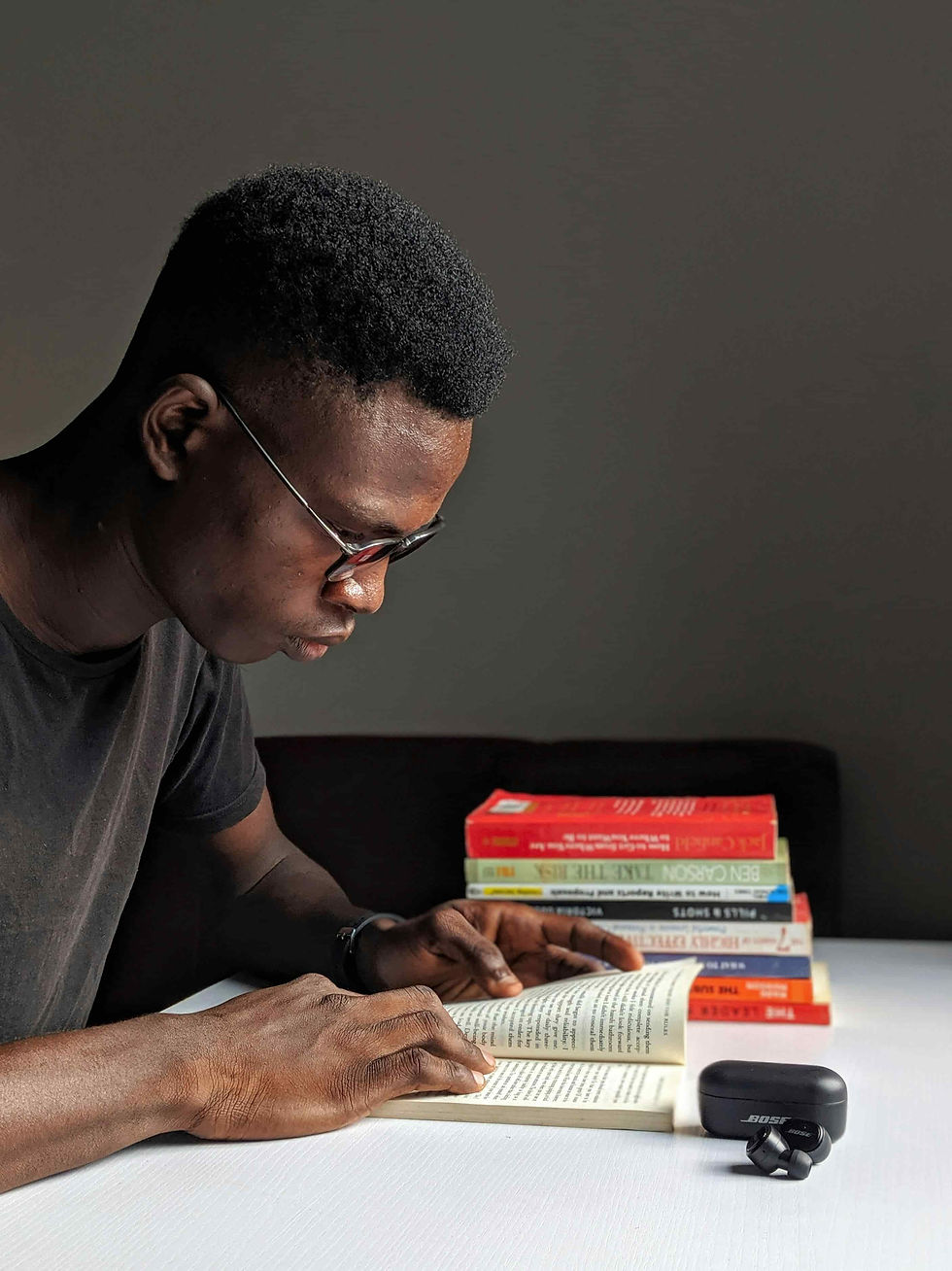 Man reading a book at a white table, stack of colorful books in the background. Dark room setting, focused and calm mood. Earbuds on table.