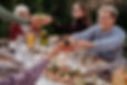 Friends sharing food at an outdoor table with colorful dishes, drinks, and candles. One person smiling, passing a bowl of fruit. Casual setting.