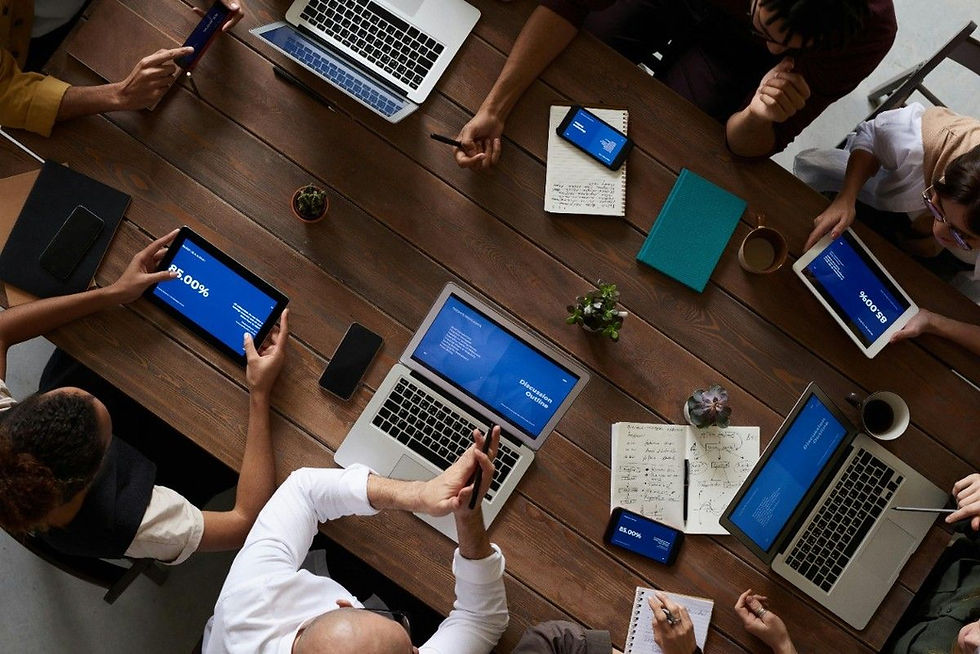 People collaborating at a wooden table with laptops, tablets, phones displaying blue screens. Papers, notebooks, and coffee cups present.