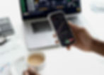 Hand holds phone displaying stock chart, other hand holds coffee. Laptop, calculator in background. White, modern, financial setting.