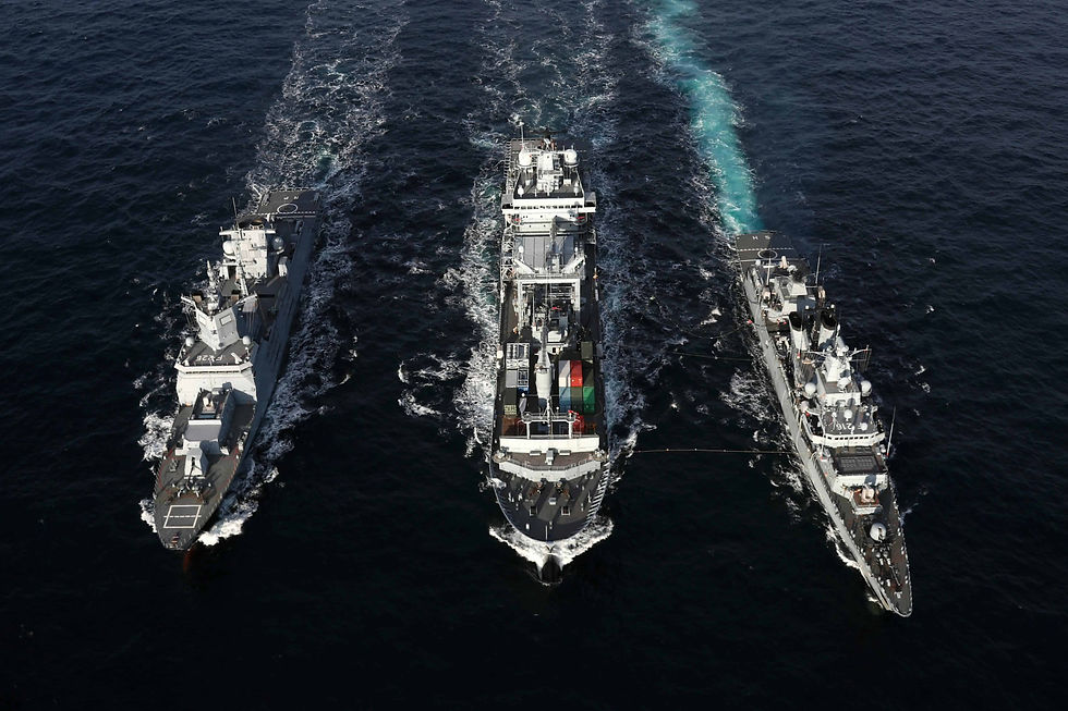 Three naval ships sail in formation on open sea, with the central one carrying containers. Calm water, deep blue sea.