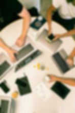 Overhead view of people working at a round table with laptops, notebooks, and coffee. A paper with a diagram is visible; relaxed vibe.