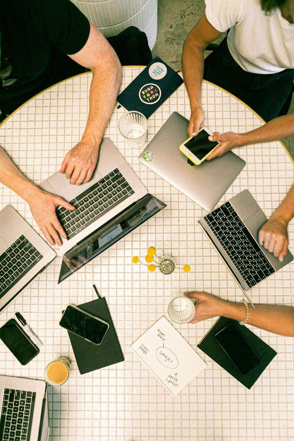 Overhead view of people working at a round table with laptops, notebooks, and coffee. A paper with a diagram is visible; relaxed vibe.
