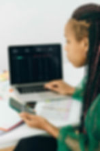 Woman with braids in green top uses a phone and laptop displaying graphs. Papers with charts are on the desk. Mood: focused.