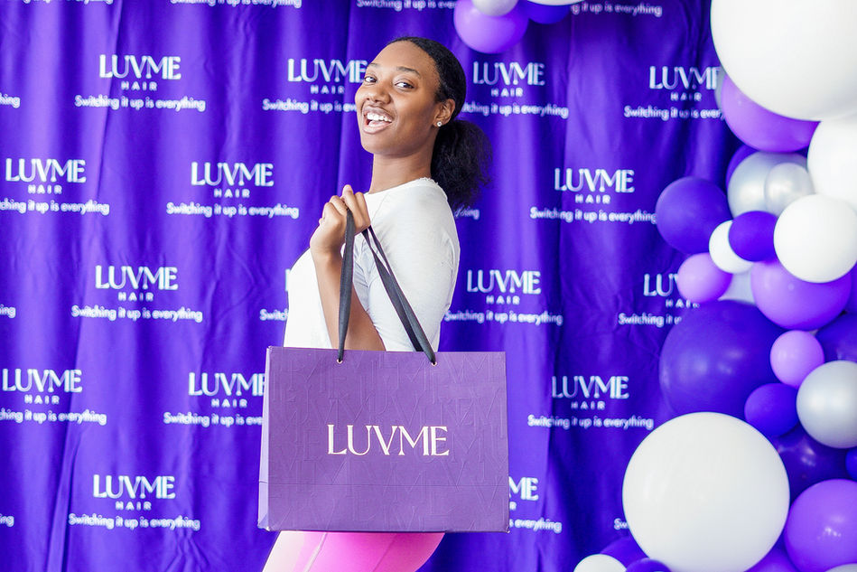 In the midst of a luvme popup hair event, a fashionable lady proudly displays a purple shopping bag against a captivating purple backdrop. Captured by starmade production.