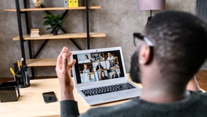 A person on a video call sits at a desk with a laptop. Shelves in the background hold books and plants, creating a calm home office setting.