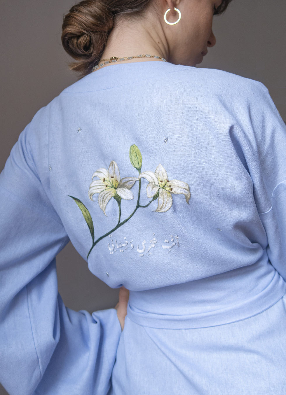 A blue linen robe adorned with hand-embroidered white lilies and Arabic calligraphy on the back, designed by Bianca Hodselle.