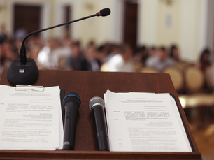 podium with microphones and printed agenda sheets facing a blurred audience in a formal hall.