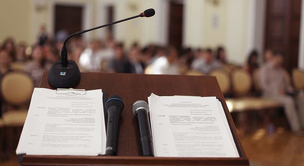 podium with microphones and printed agenda sheets facing a blurred audience in a formal hall.