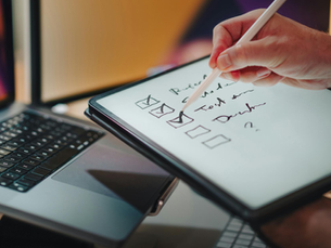 hand using a stylus on a tablet checklist beside a laptop in a warm-lit workspace.