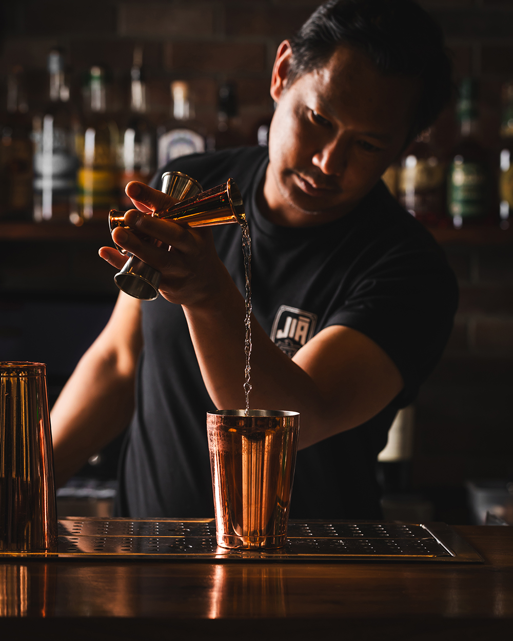 “Bartender mixing a drink behind the bar, low light.”