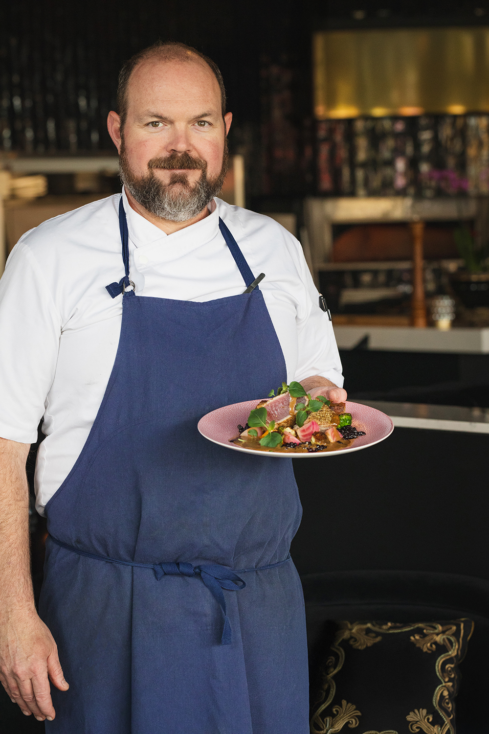 “Chef in apron holding a plated dish, restaurant portrait.”