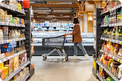 Female grocery shopper with cart browsing refrigerated section in supermarket aisle, illustrating grocery product information management and retail shopping experience.