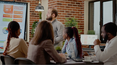A diverse group of five business professionals gathered around a conference table, listening to a man presenting in front of a large screen. The screen displays an 'ANALYTICS SUMMARY' with charts and data, including a prominent '25:1' ratio. The setting appears to be a modern office or meeting room with exposed brick walls. This image represents collaborative business analytics and decision-making, possibly facilitated by a unified platform