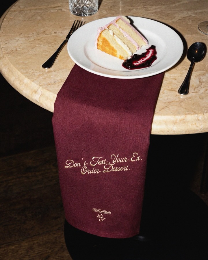 A slice of layered cake with berry compote served on a marble table featuring a burgundy napkin embroidered with "Don't Text Your Ex. Order Dessert."