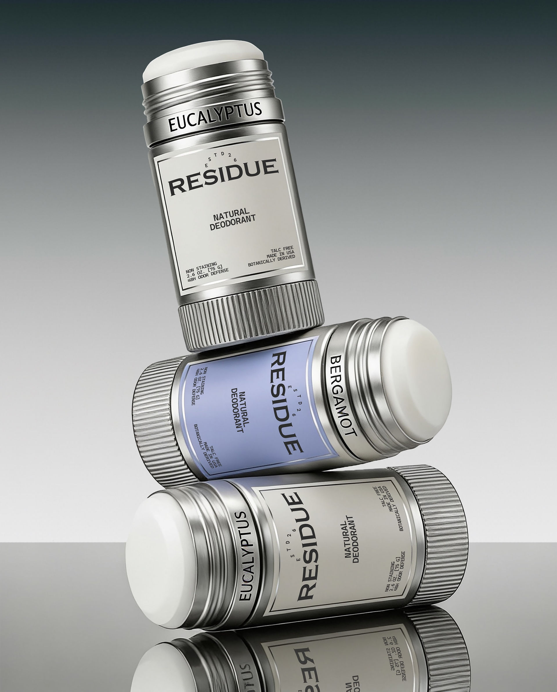 Three natural deodorant sticks stacked diagonally, showcasing silver packaging, white and powder blue labels, exposed deodorant formula, and engraved fragrance names.
