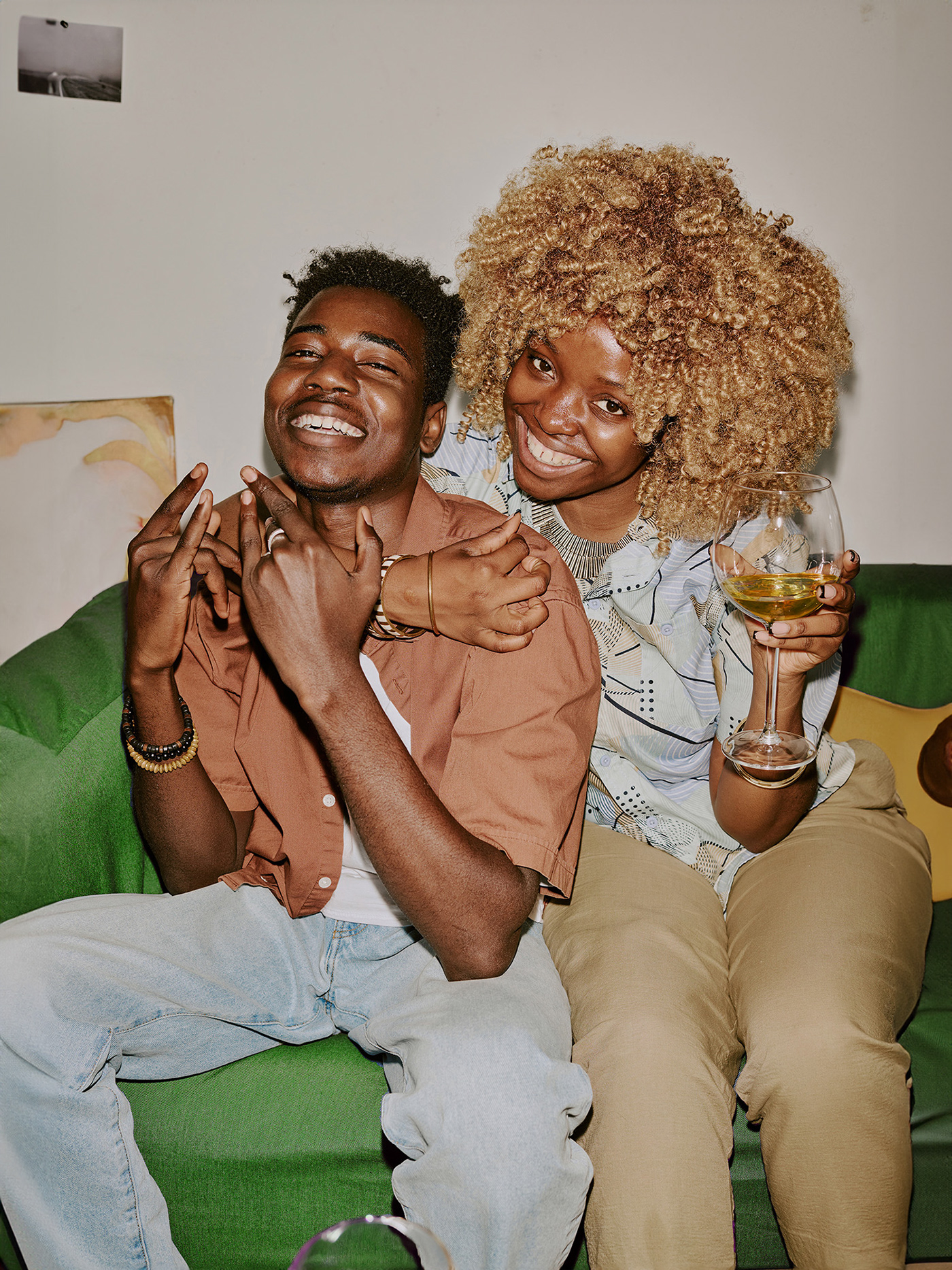 Lifestyle portrait of two friends sitting on a green couch, smiling and holding wine glasses, representing Luminéa’s warm, modern, community-driven brand aesthetic.