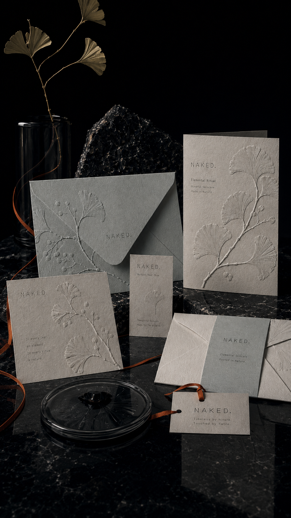 NAKED. brand identity stationery with embossed botanical cards, envelope, hang tag, and printed collateral arranged on dark marble with subtle amber ribbon accents.