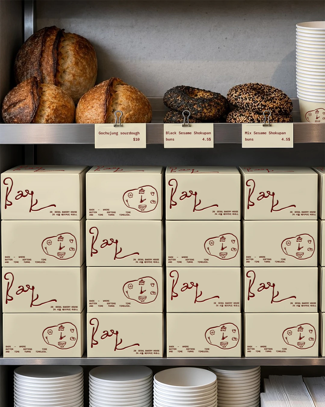 Baek Korean Bakery display shelf with cream branded pastry boxes stacked beneath fresh bread, sesame buns, product labels, cups, plates, and warm bakery packaging details.