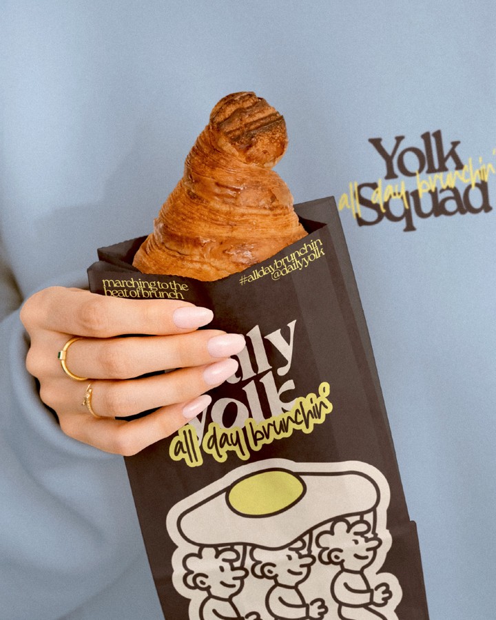Daily Yolk brunch café branding featuring a croissant in a custom illustrated takeaway bag with bold typography and playful egg character artwork.