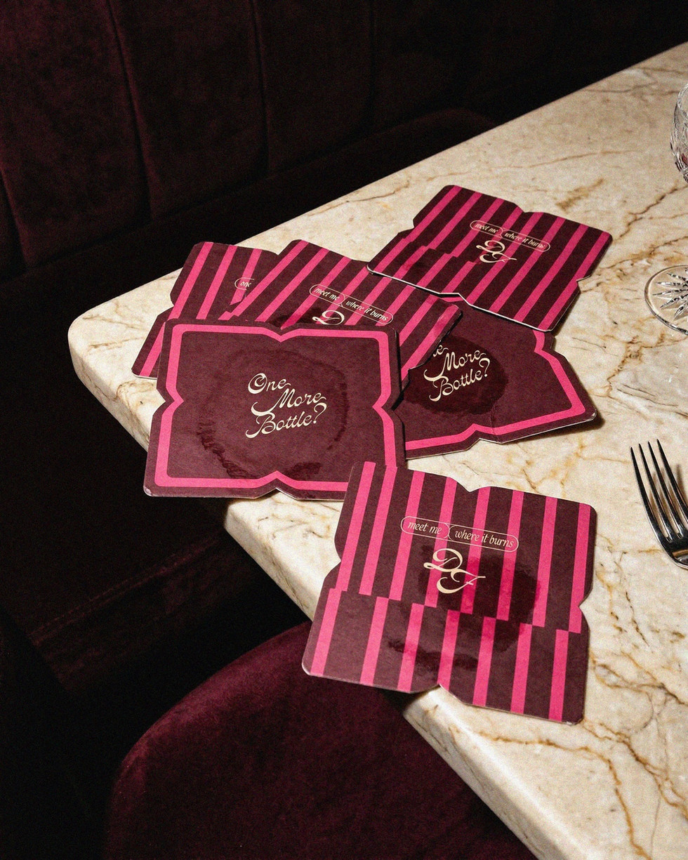 A scattered pile of burgundy and pink striped coasters on a marble tabletop, featuring the playful script "One More Bottle?".