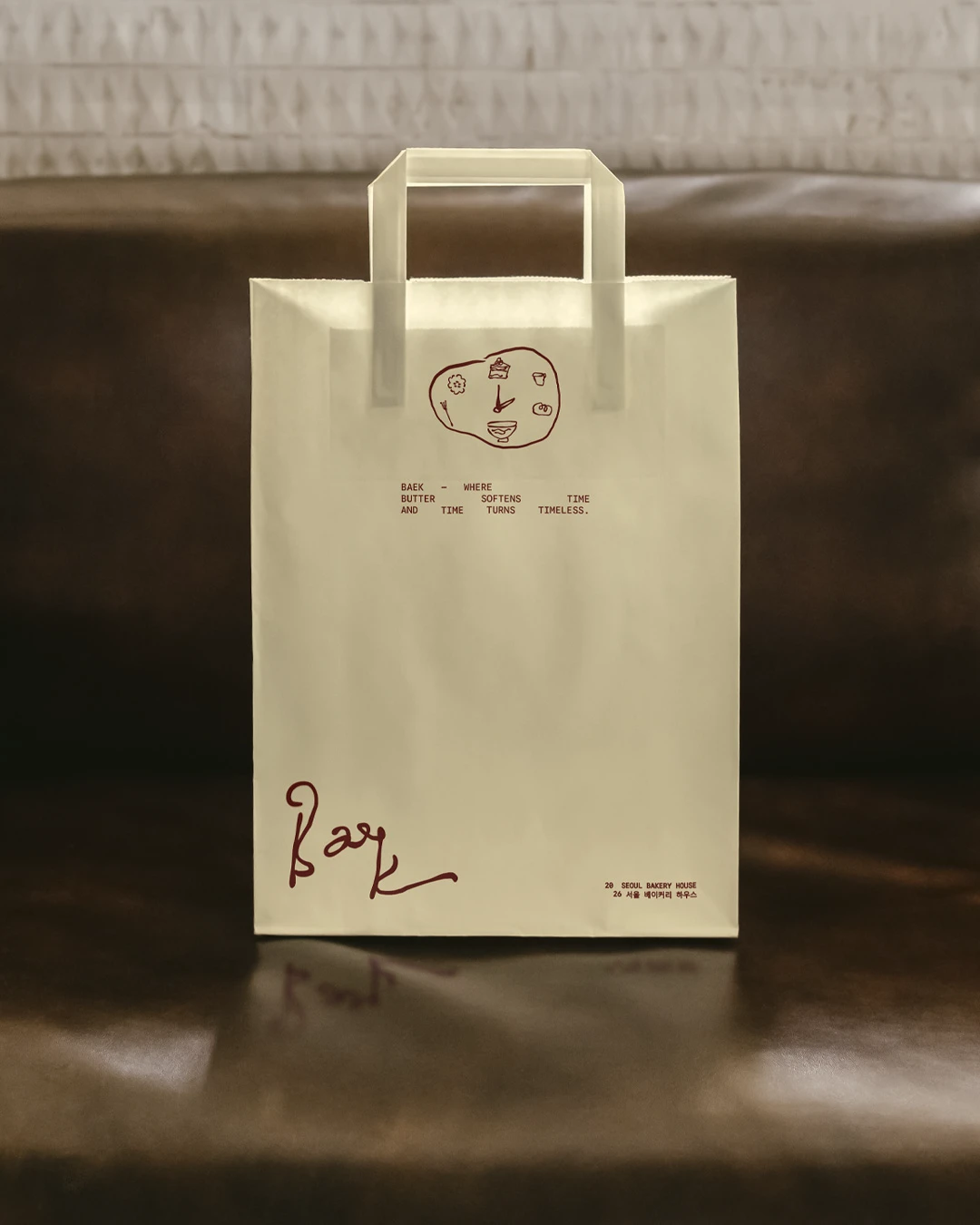Baek Korean Bakery cream paper shopping bag mockup on a dark leather seat, featuring deep red handwritten logo details, small bakery illustrations, Korean text, and a minimal tagline.