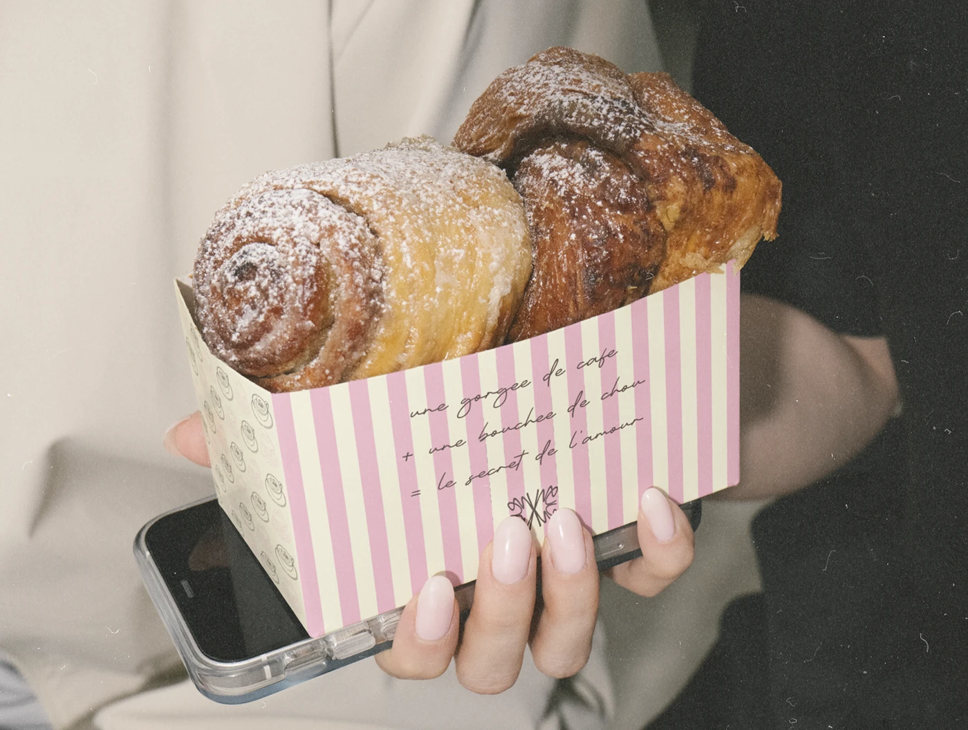 Hand holding a striped pink-and-cream Mon Chou pastry box filled with powdered rolls, showcasing the bakery’s soft pastel packaging and handwritten French details.
