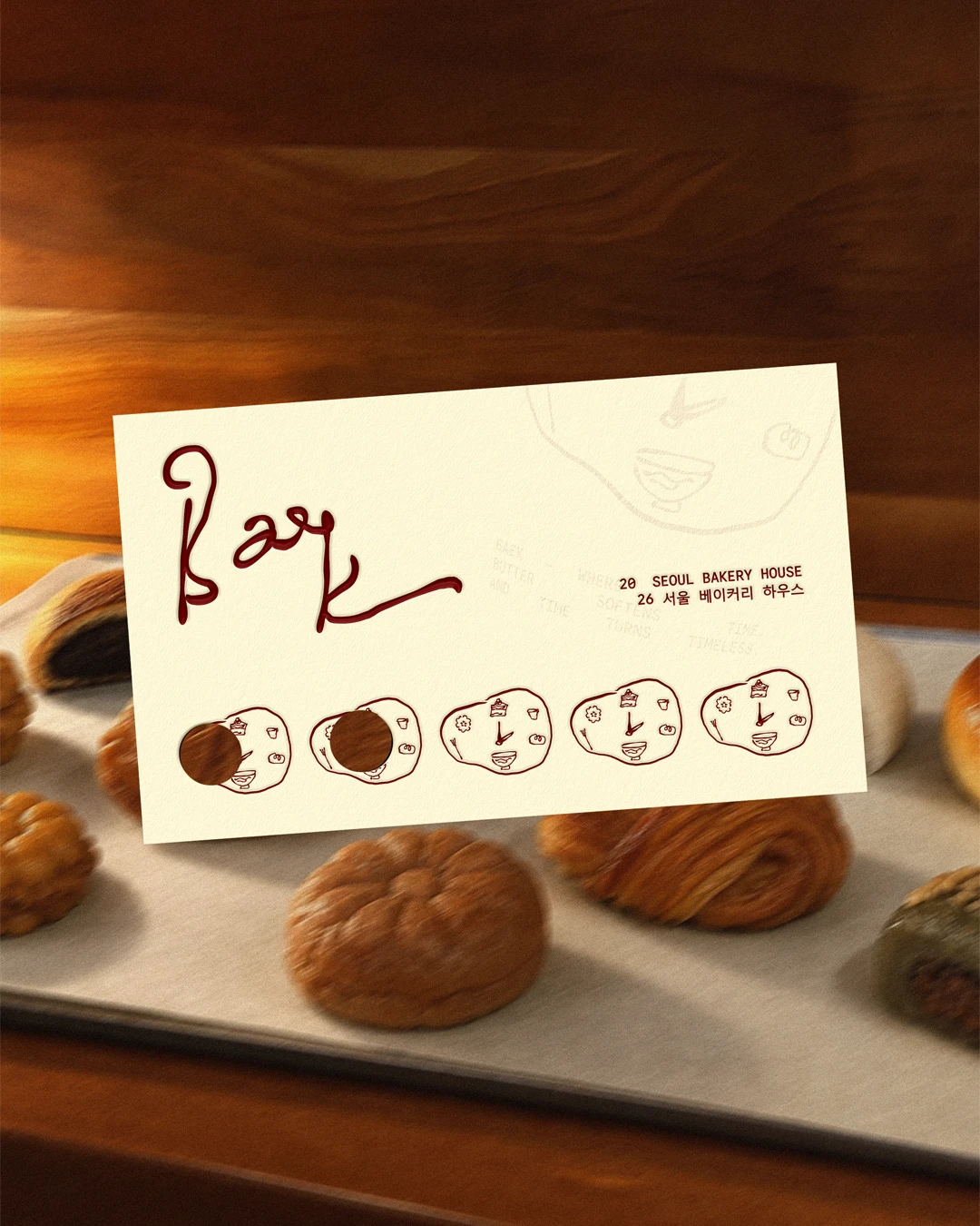 Baek Korean Bakery printed brand card shown above a tray of pastries, featuring the deep red handwritten logo, small illustrated icon pattern, and warm bakery photography.