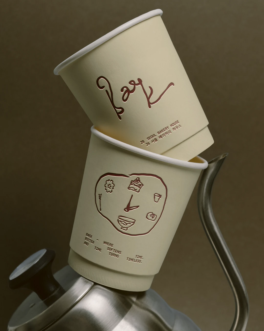 Baek Korean Bakery branded paper cups stacked on a metal kettle, featuring cream packaging, deep red logo marks, small bakery illustrations, and Korean text.
