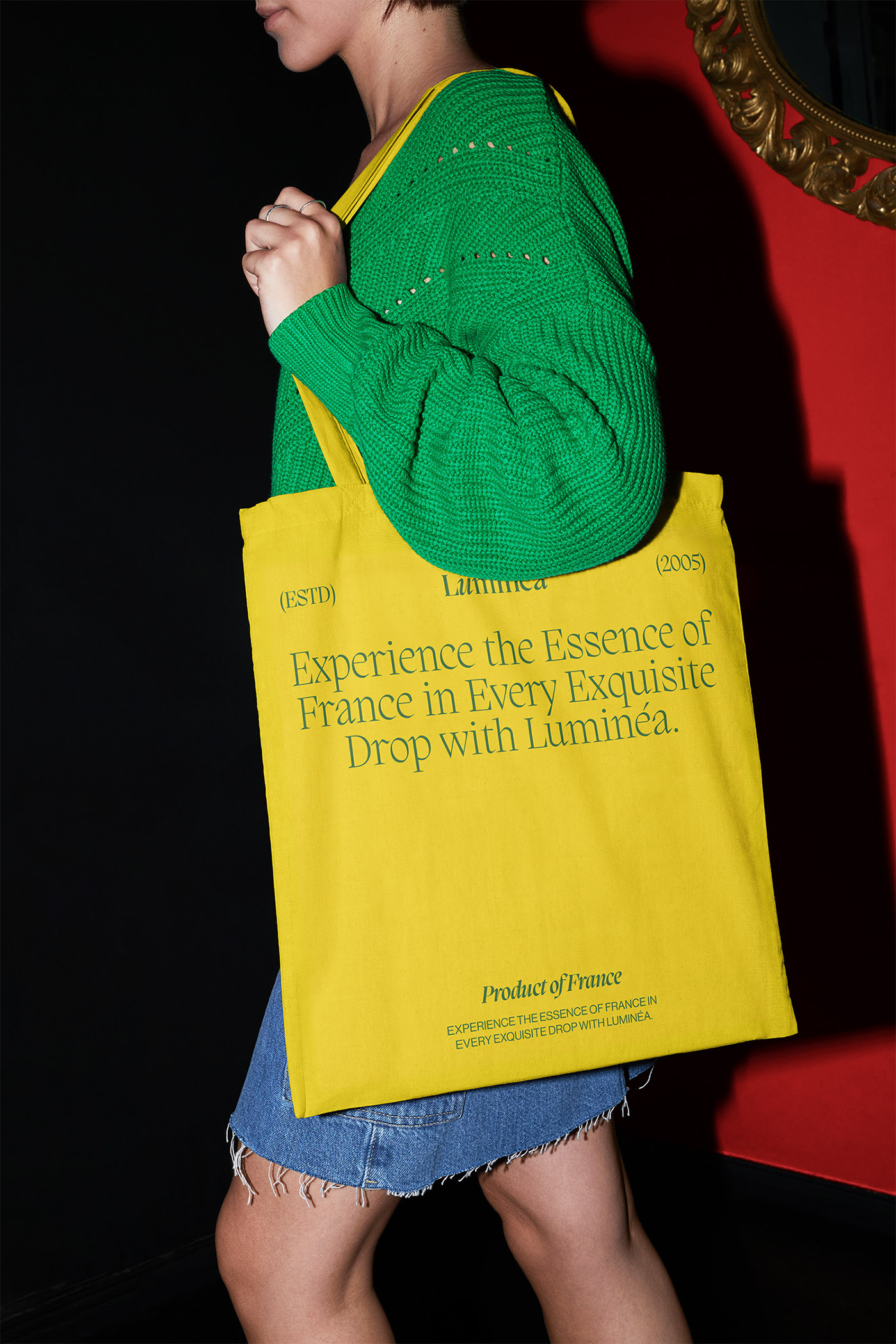 Person wearing a green sweater and denim skirt carrying a bright yellow Luminéa tote bag featuring large editorial typography and bold brand styling.