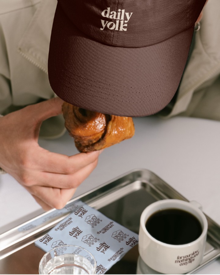 Daily Yolk brunch café brand application showing logo-embroidered cap, branded coffee cup, illustrated paper goods, and pastry on a café tray.